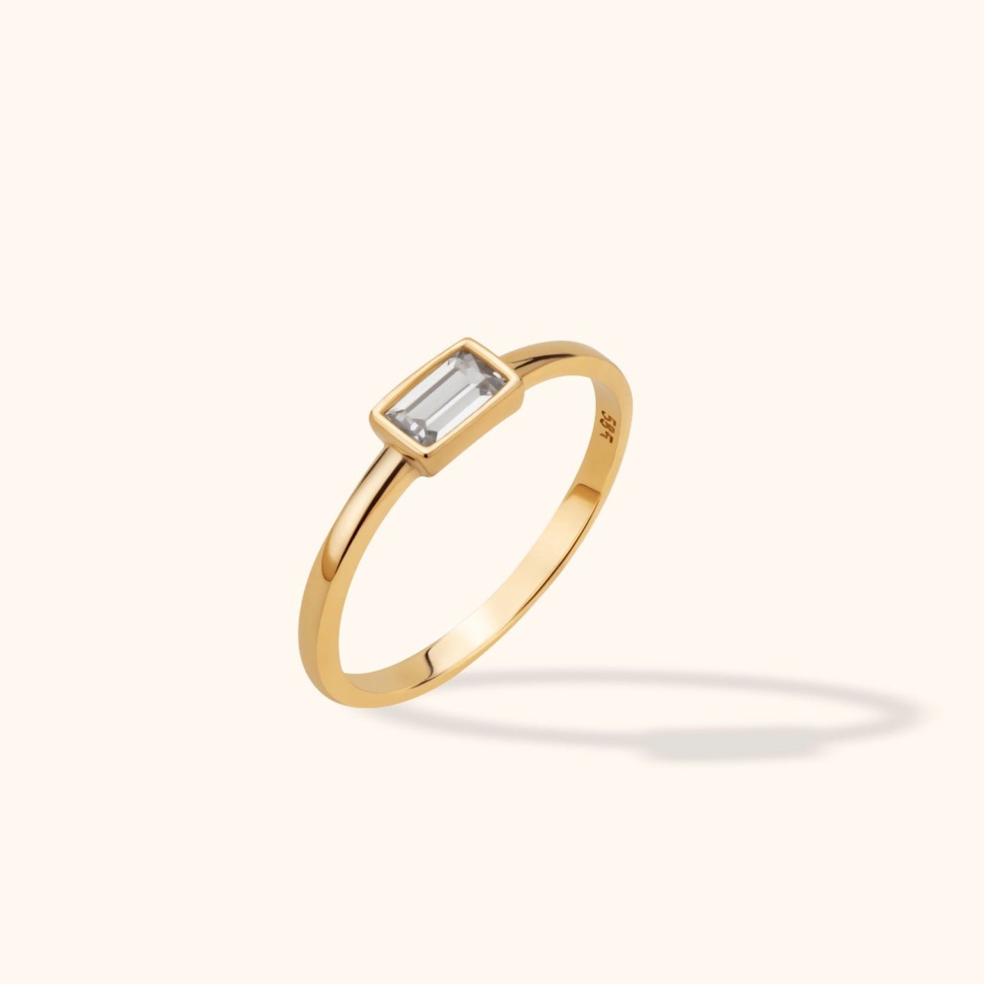 Slim gold-toned ring with a clear rectangular stone in a bezel setting, shown at an angle on a pale cream background.