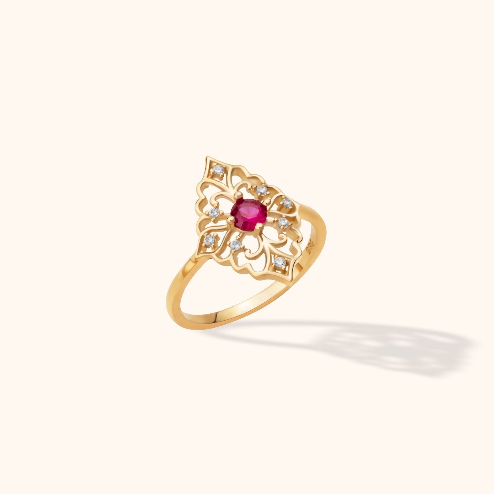 Gold-toned ring with an ornate openwork floral face, a round deep pink center stone, and small clear accents.