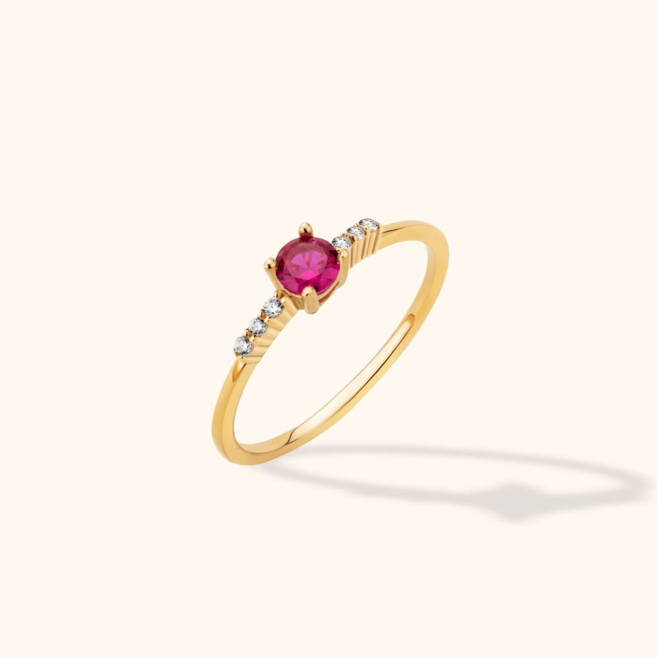 Slim gold-colored ring with a round pink center stone and three small clear stones on ridged shoulders on each side.