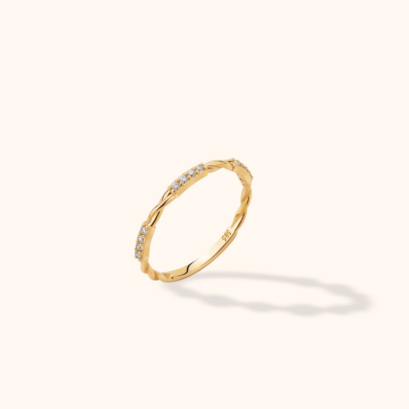 Slim gold-colored ring with twisted sections alternating with rows of small clear stones, shown tilted on a cream background.