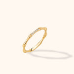 Slim gold-colored ring with bamboo-like segments and a row of small clear stones, tilted against a cream background.