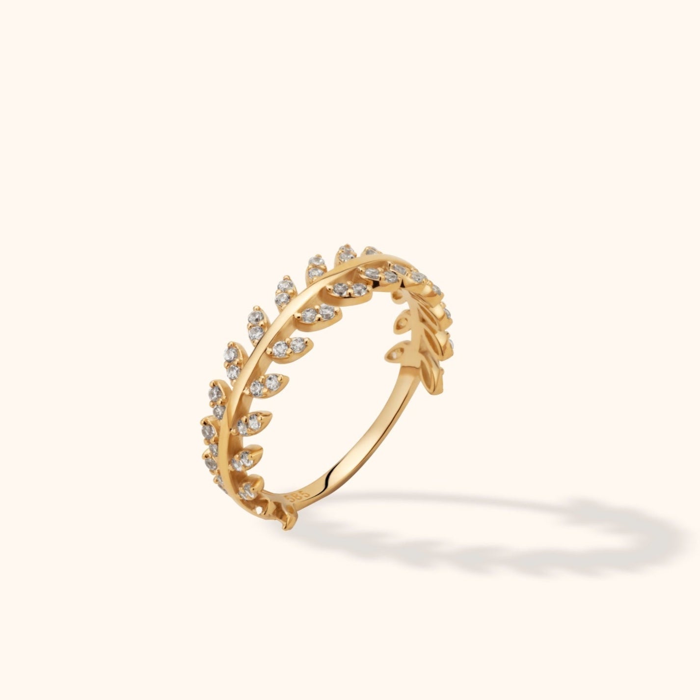 Gold-colored ring with leaf-shaped details and small clear stones, shown tilted on a cream background with a soft shadow.