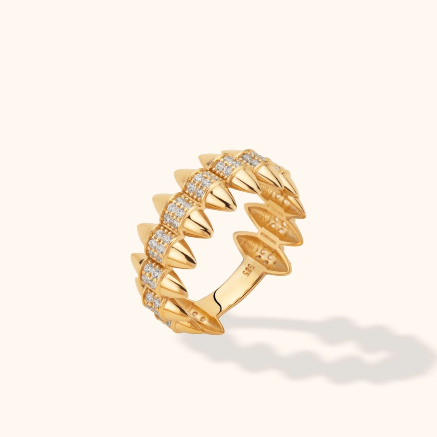 Gold-colored ring with polished spikes along both edges and rows of small clear stones, shown at an angle on a cream background.