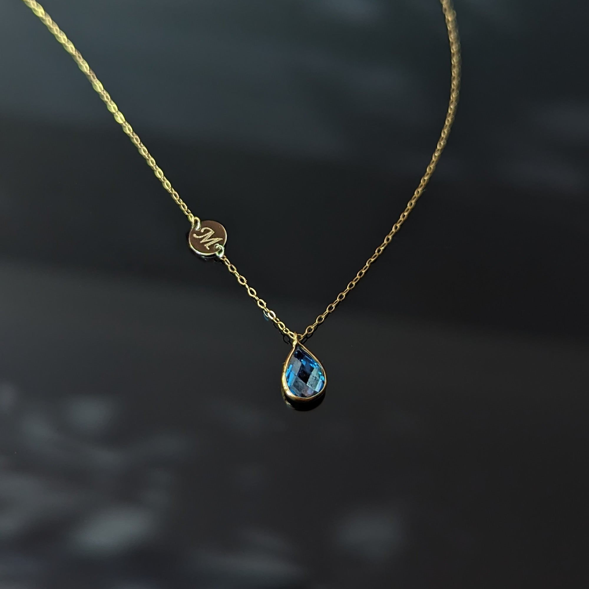 Gold-toned chain necklace with a faceted blue teardrop pendant and a small round disc marked M, against a dark background.