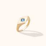 Gold-toned signet ring with a square face and a round blue faceted center in a star-shaped setting, shown at an angle.