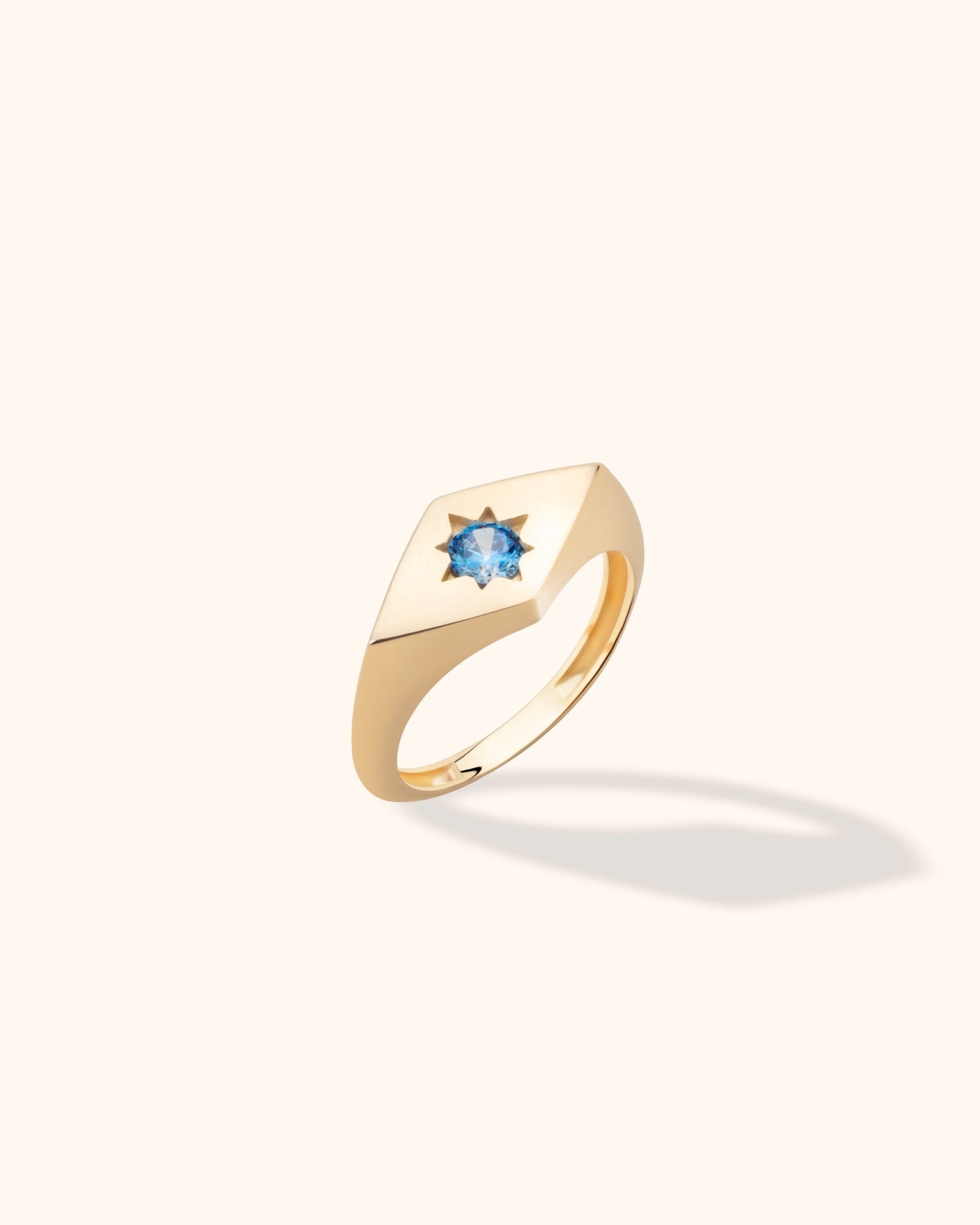 Gold-toned signet ring with a square face and a round blue faceted center in a star-shaped setting, shown at an angle.