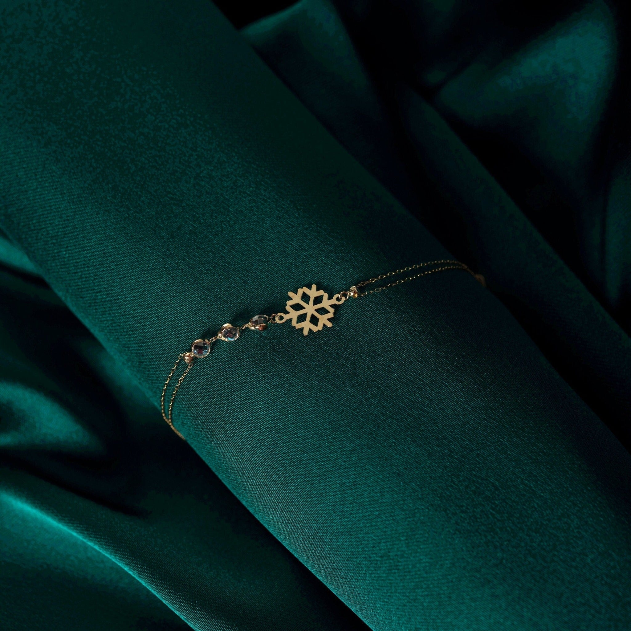 Gold-tone bracelet with a cutout snowflake charm, fine double chains and three clear faceted accents on dark green fabric.
