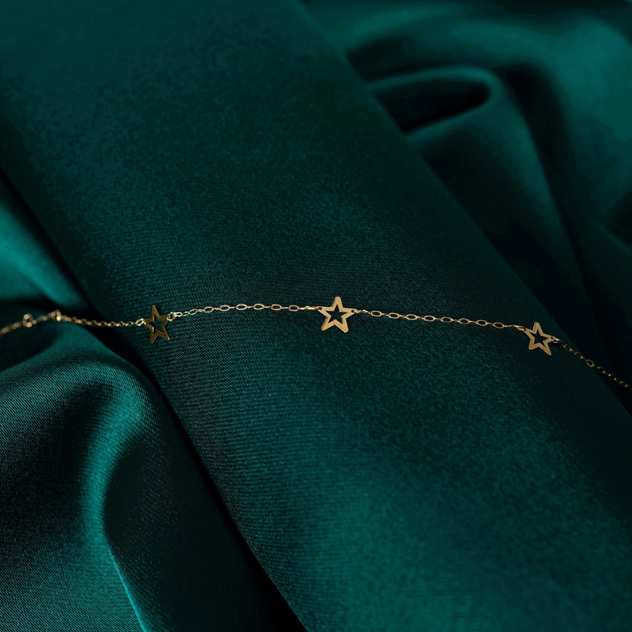 Gold-toned fine chain bracelet with three open star shapes, laid across folds of dark teal fabric.