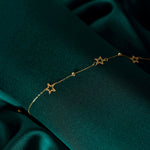 Fine gold-toned chain bracelet with open star shapes and small textured beads, draped diagonally over dark teal fabric.
