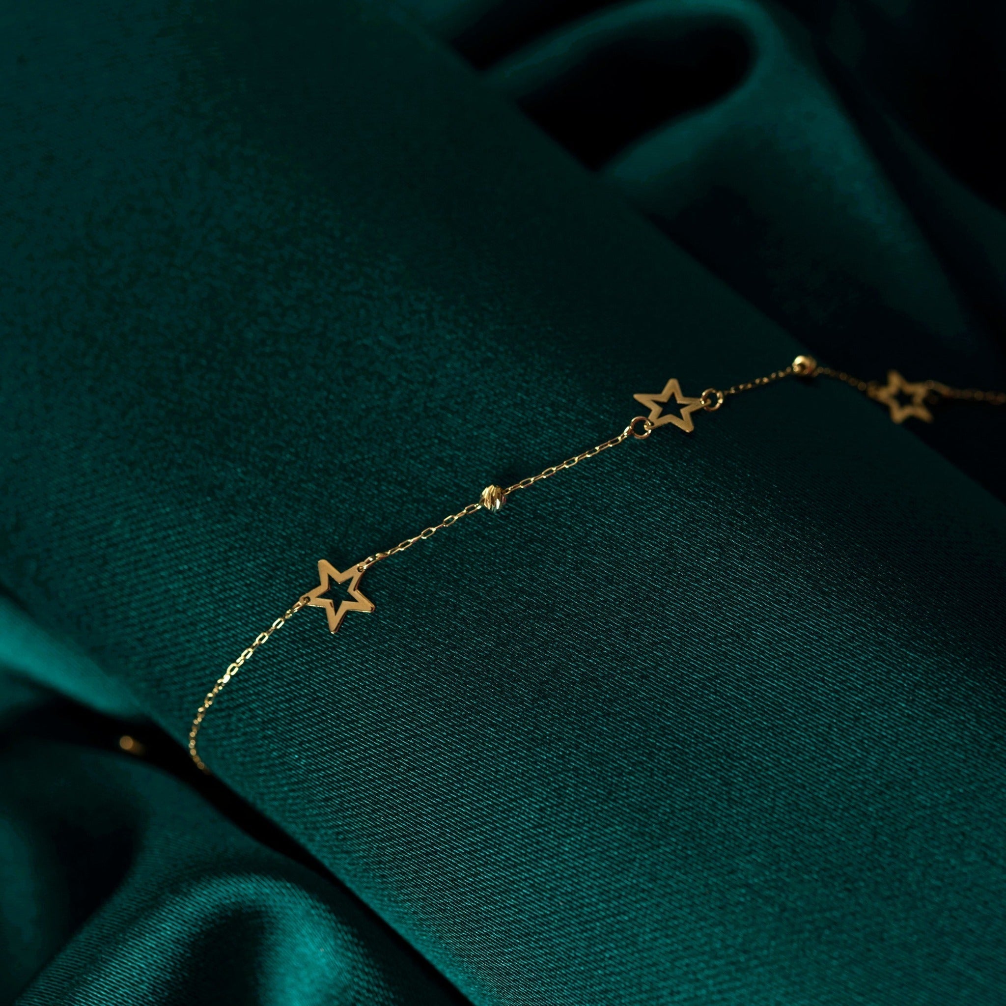 Fine gold-toned chain bracelet with open star shapes and small textured beads, draped diagonally over dark teal fabric.
