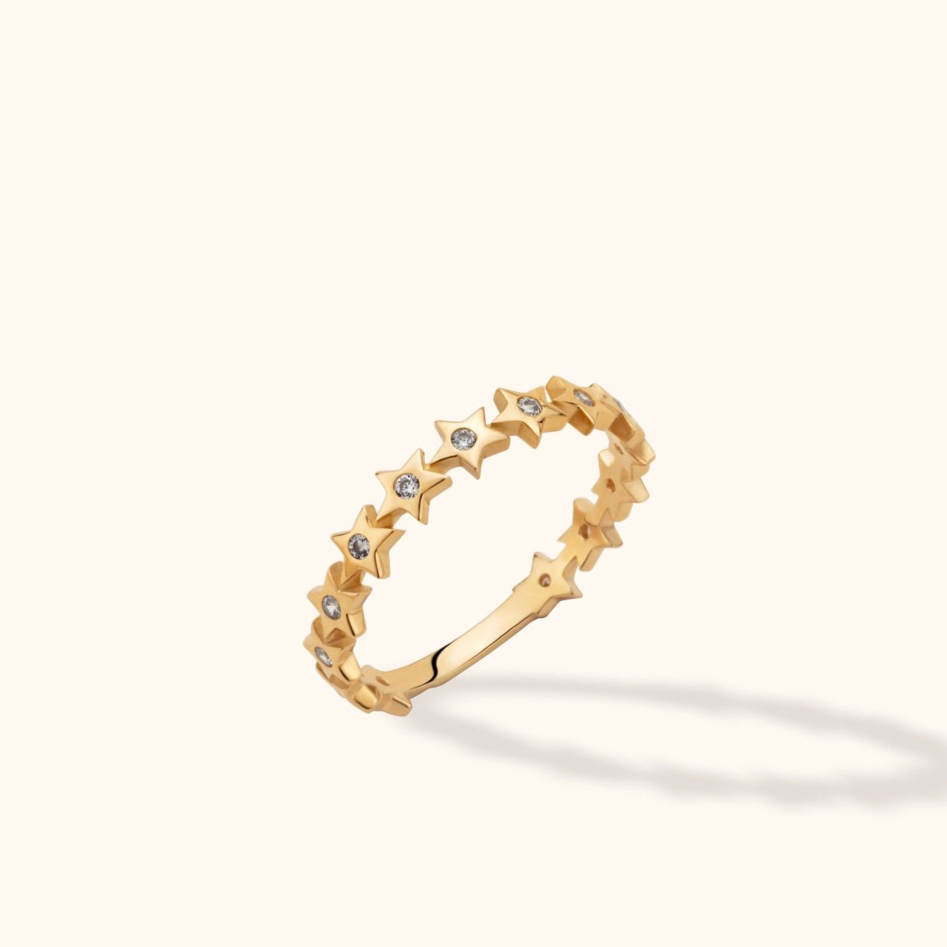 Gold-colored ring with a row of five-pointed stars and small clear round accents, tilted against a cream background.
