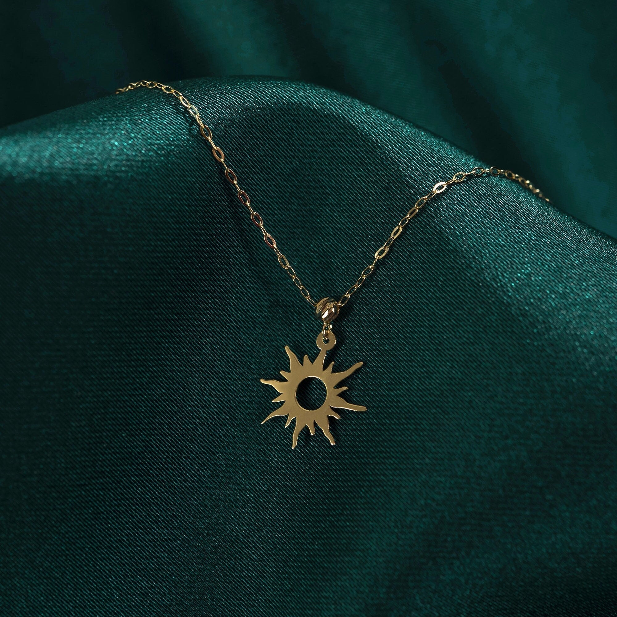 Gold-colored oval-link necklace with a sunburst pendant and circular cutout center, resting on dark green fabric.