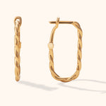 Pair of gold-colored twisted hoop earrings with elongated, rounded rectangular outlines, shown from two angles on a cream background.