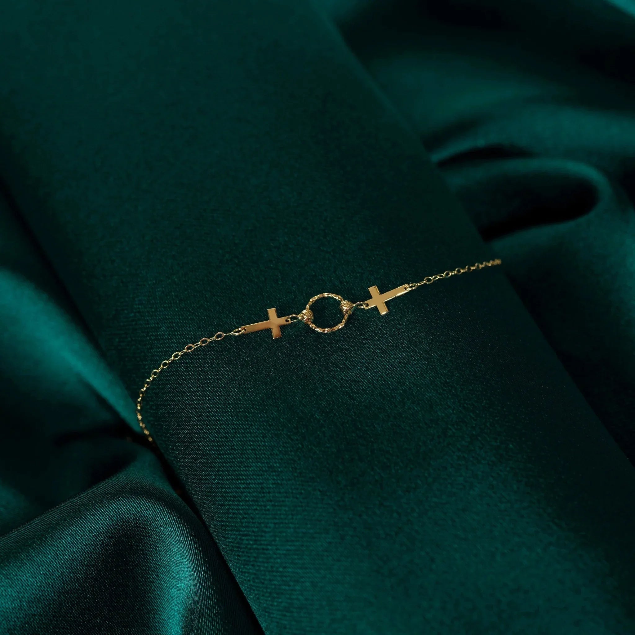 14K gold bracelet with two cross charms and a textured circular centerpiece on deep green fabric.