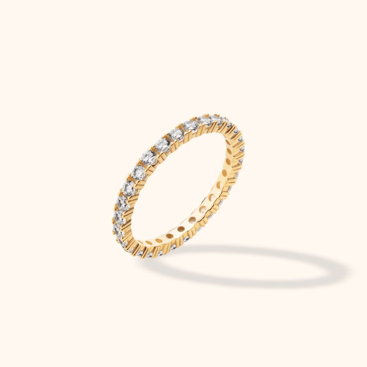 Gold-toned ring with a continuous row of round clear stones and circular openings inside the band, tilted on a cream background.