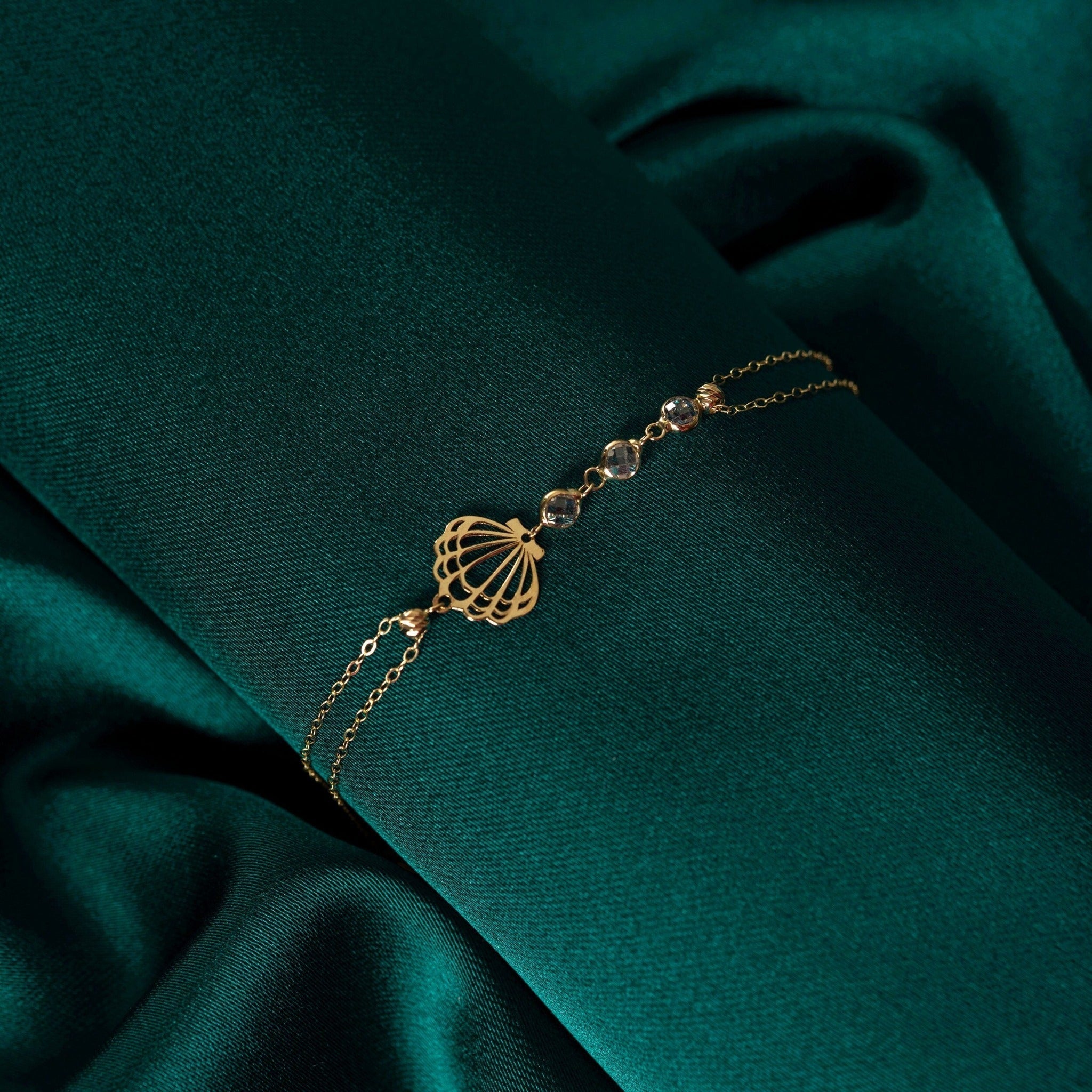 Gold-tone double-chain bracelet with an openwork seashell charm and three clear faceted beads on dark teal fabric.