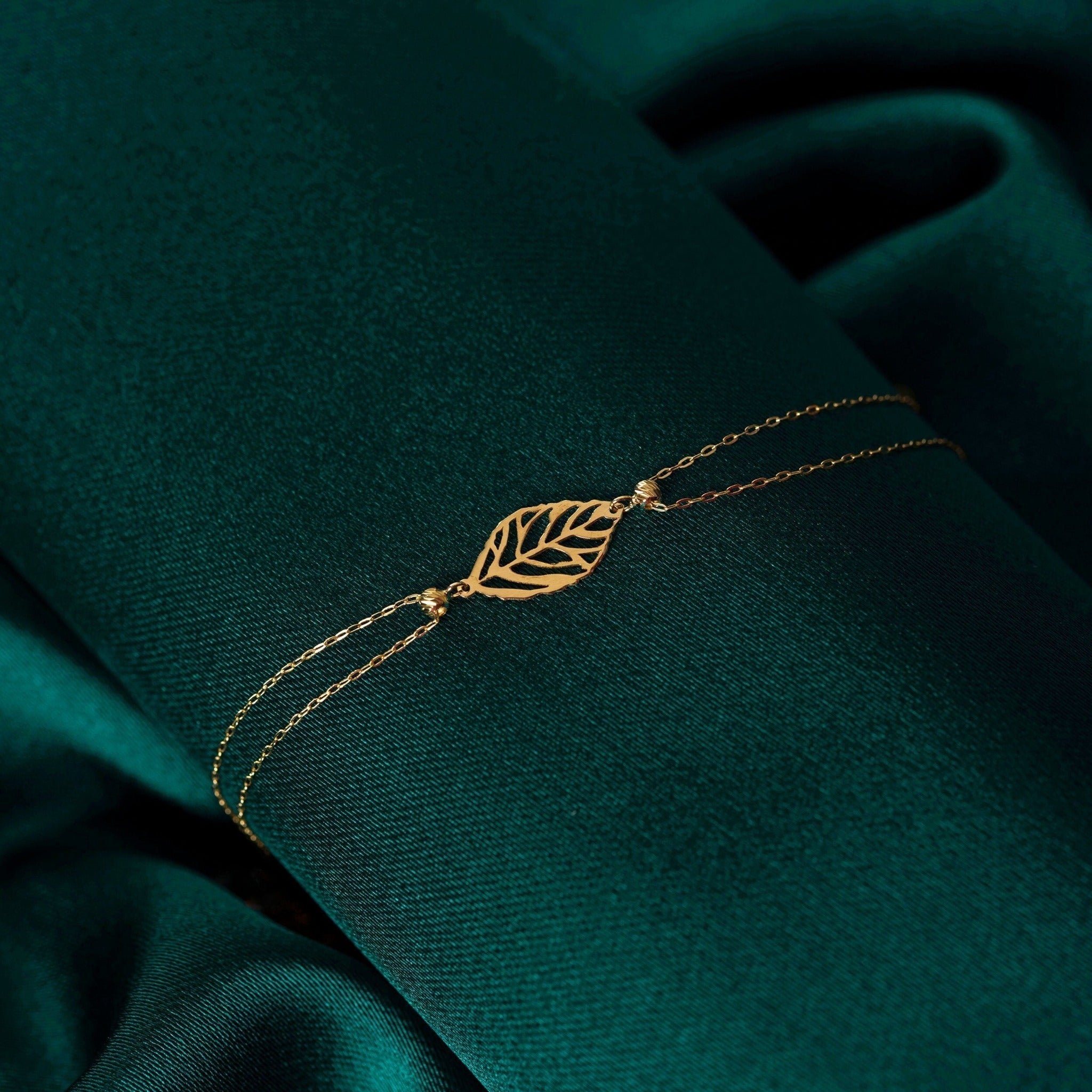 Gold-tone bracelet with an openwork leaf centerpiece, double chains and small beads, resting on dark teal fabric.