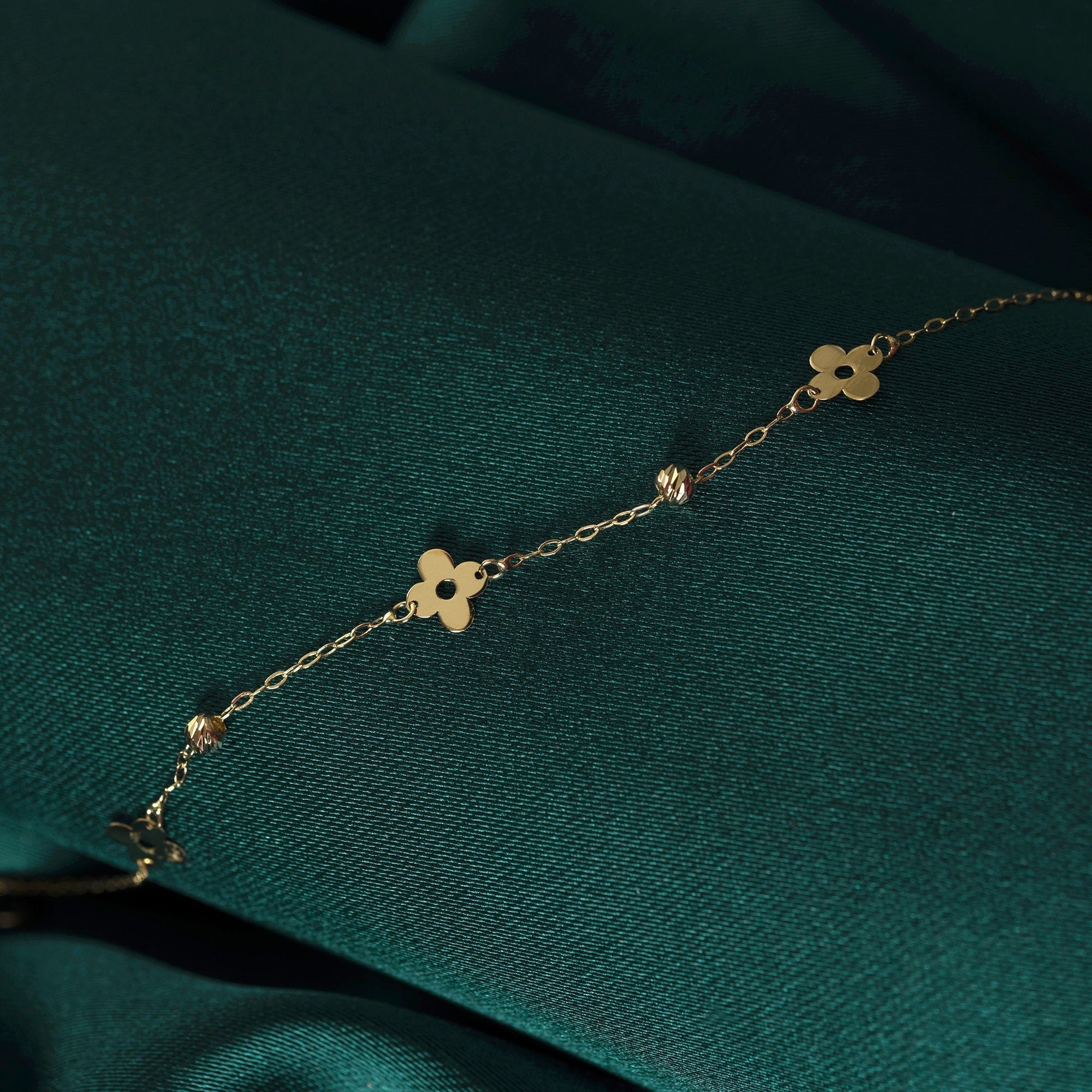 Gold-toned chain bracelet with flat clover shapes, circular cutouts and faceted beads, laid diagonally on dark green fabric.