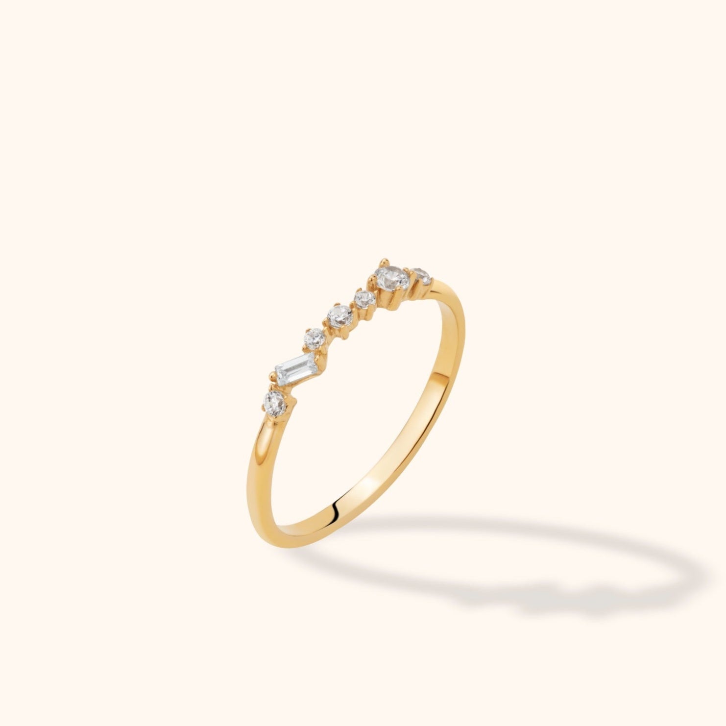 Slim gold-toned ring with an asymmetric row of clear round stones and one rectangular stone, shown at an angle on a cream background.