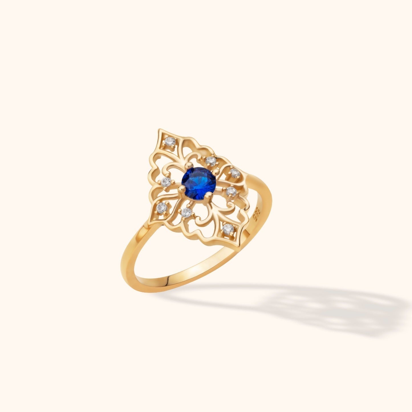 Gold-toned ring with an ornate openwork face, a round blue center stone, and small clear accent stones on a cream background.
