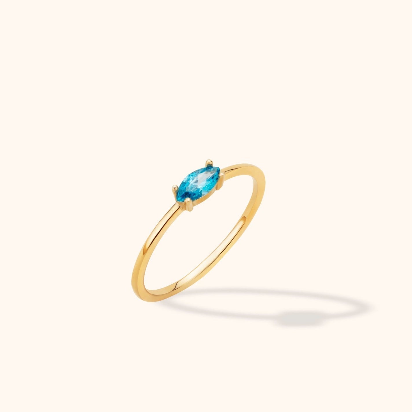 Slim gold-toned ring with a marquise-shaped blue stone in a prong setting, shown at an angle on a cream background.
