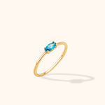 Slim gold-toned ring with a marquise-shaped blue stone in a prong setting, shown at an angle on a cream background.