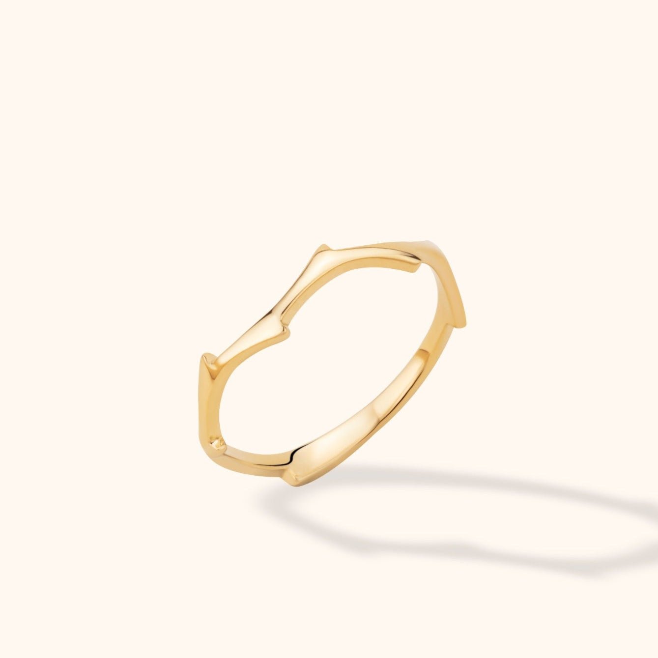 Polished gold-tone ring with angular peaks and curved edges, shown tilted against a cream background with a soft shadow.