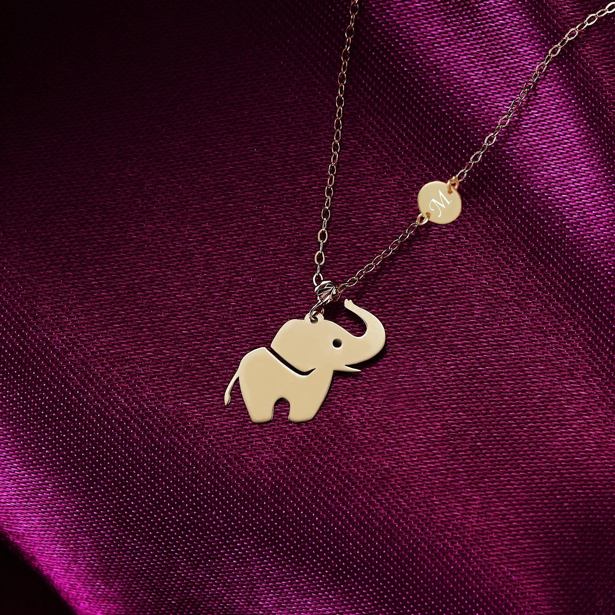 Gold-toned chain necklace with a raised-trunk elephant pendant and round M initial charm on deep purple fabric.