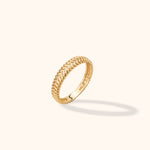 Gold-toned ring with a raised braided pattern and smooth inner band, tilted upright on a cream background with a soft shadow.