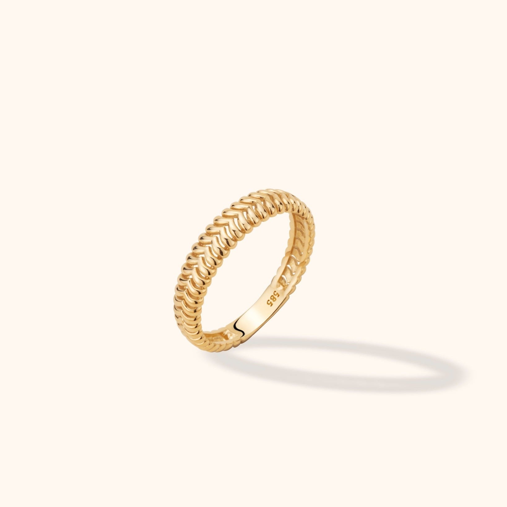 Gold-toned ring with a raised braided pattern and smooth inner band, tilted upright on a cream background with a soft shadow.