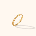 Slim gold-toned ring with an interlocking braided pattern, tilted upright on a cream background with a soft shadow.