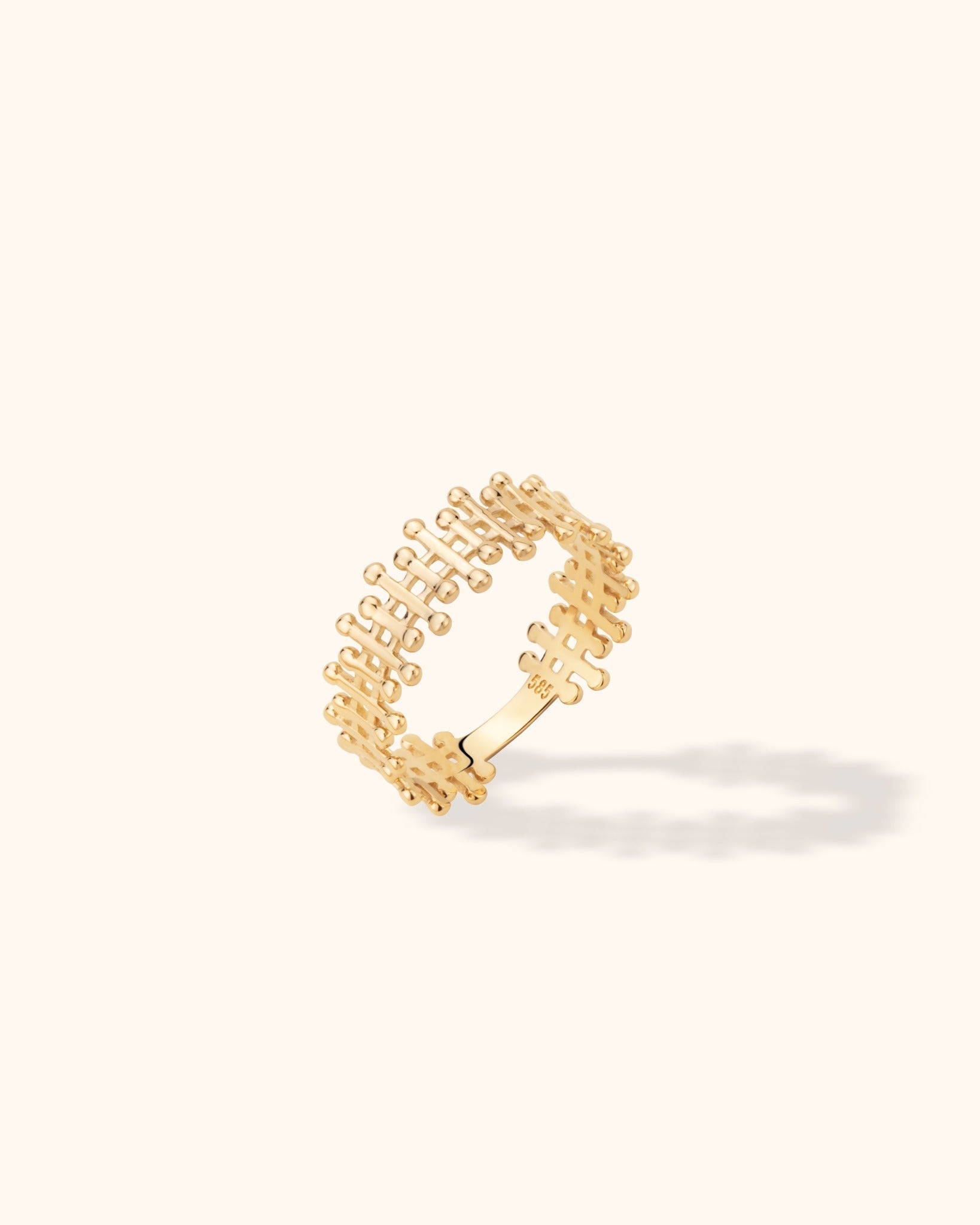 Gold-colored ring with an open grid pattern and rounded bar ends, shown tilted on a cream background with a soft shadow.