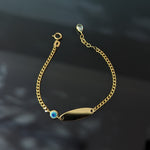Gold-tone chain bracelet with a blank oval ID plate, blue evil eye detail, round clasp and extension chain on a dark surface.