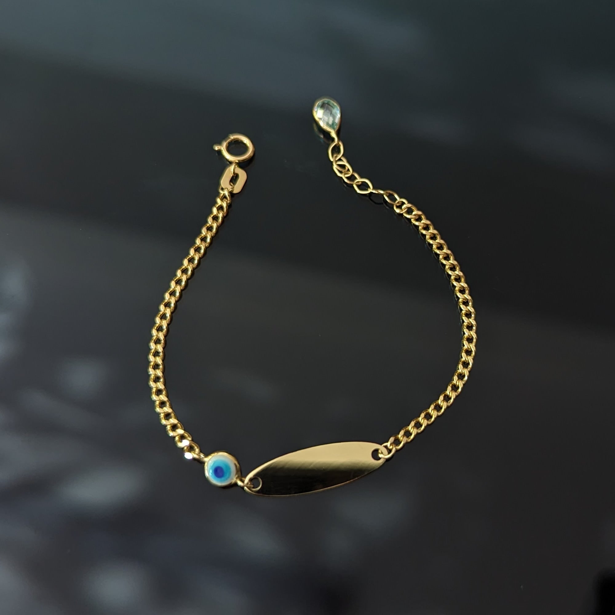 Gold-tone chain bracelet with a blank oval ID plate, blue evil eye detail, round clasp and extension chain on a dark surface.