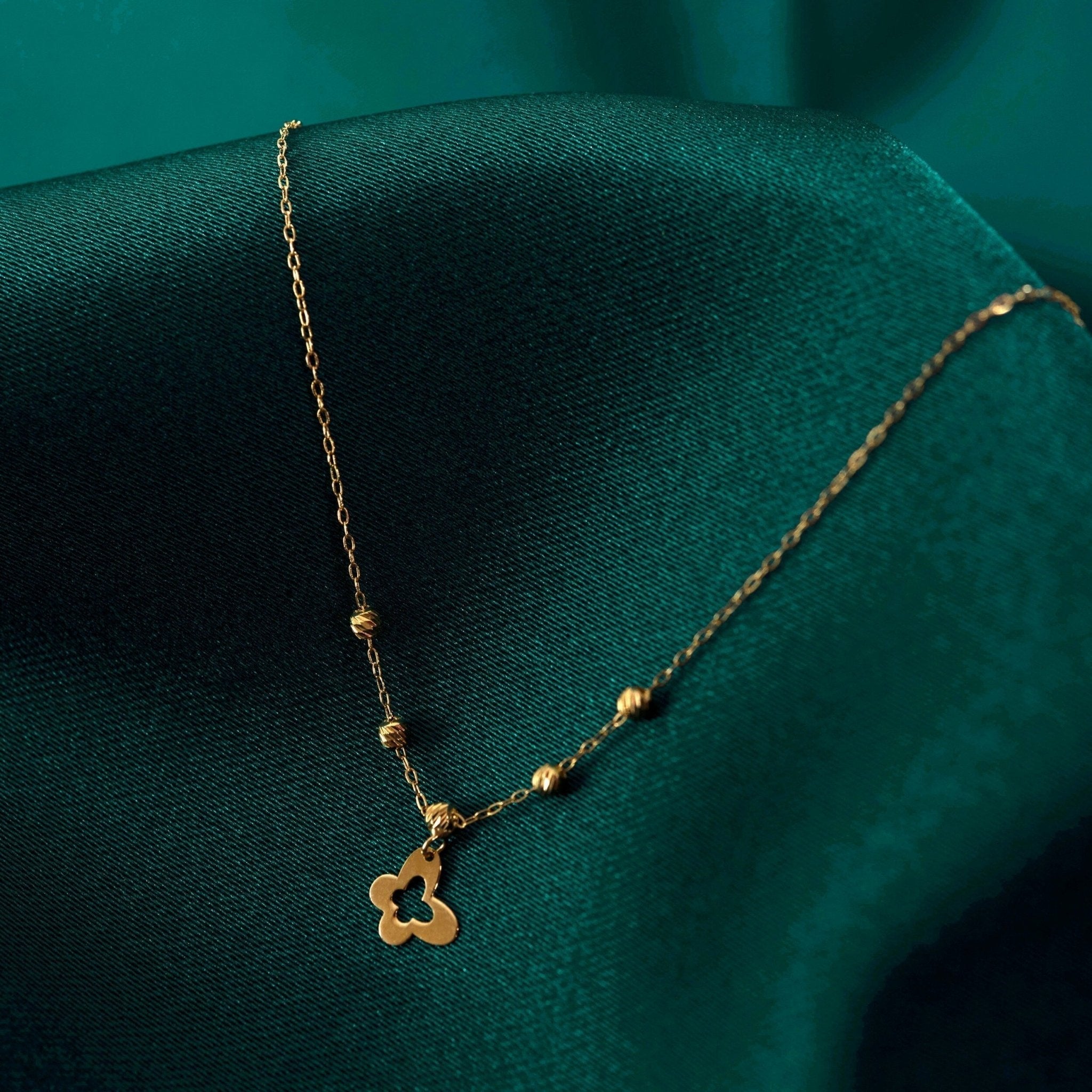 Gold-toned chain necklace with textured round beads and a cutout clover-shaped pendant on dark teal fabric.
