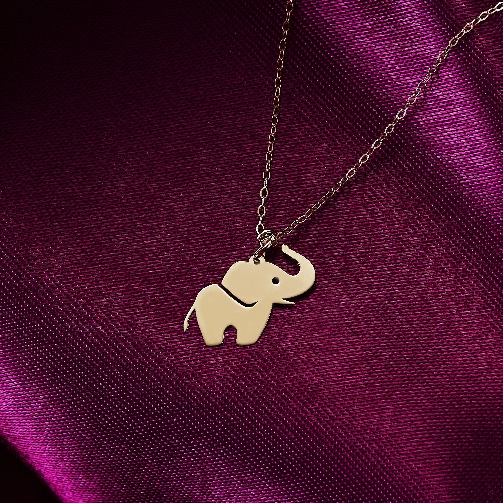 Gold-tone necklace with a flat elephant pendant, raised trunk and cutout eye, resting on glossy purple fabric.