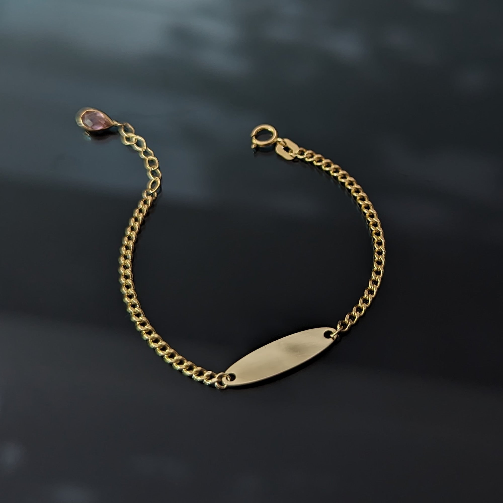 Gold-tone chain bracelet with a blank oval ID plate, pink teardrop charm, extension links and a round clasp on a dark surface.