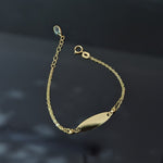 Gold-toned chain bracelet with a blank oval nameplate, round clasp, extension links and a pale blue teardrop accent on a dark background.