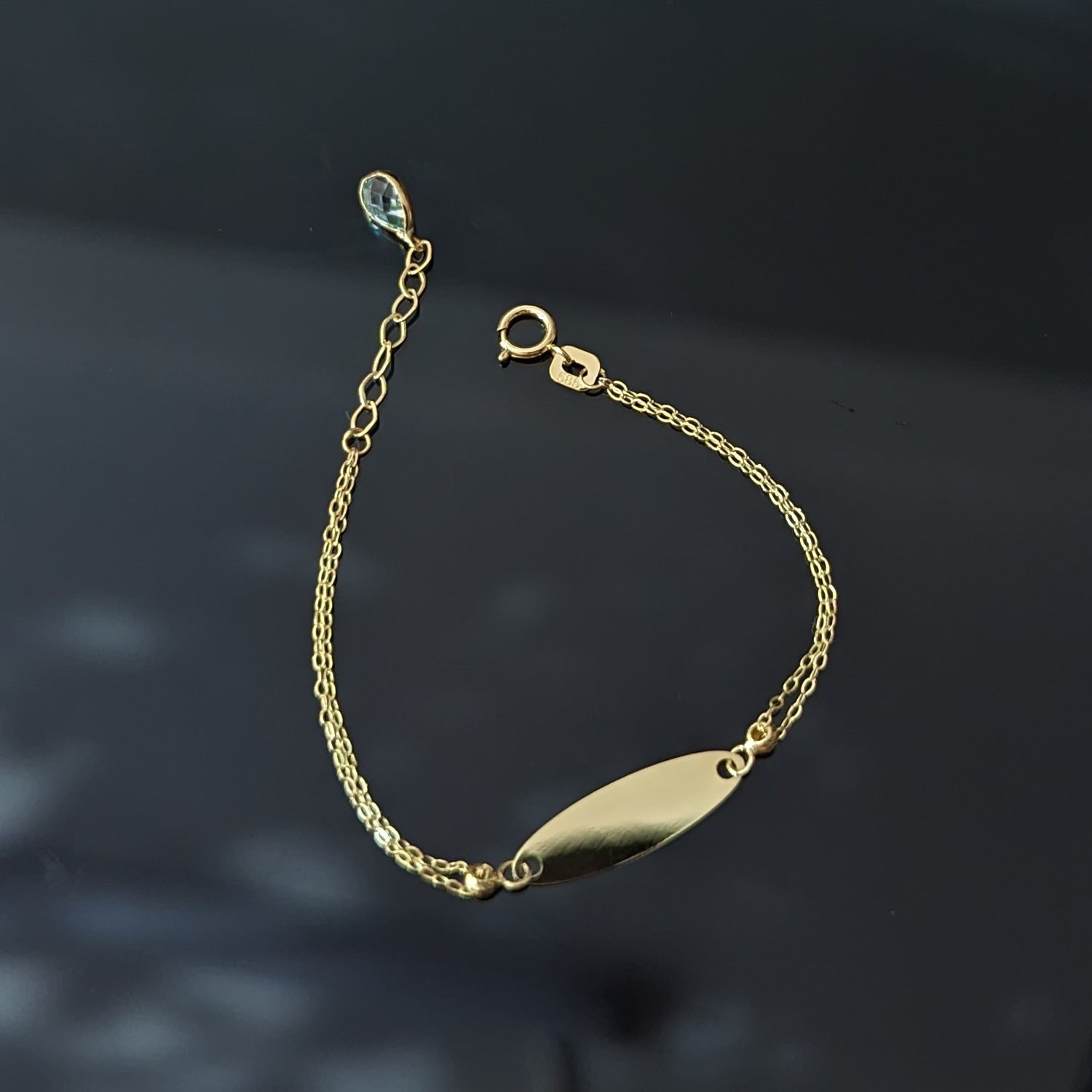 Gold-toned chain bracelet with a blank oval nameplate, round clasp, extension links and a pale blue teardrop accent on a dark background.