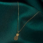 Gold-colored necklace with a fine oval-link chain and open lattice pineapple pendant on dark teal fabric.