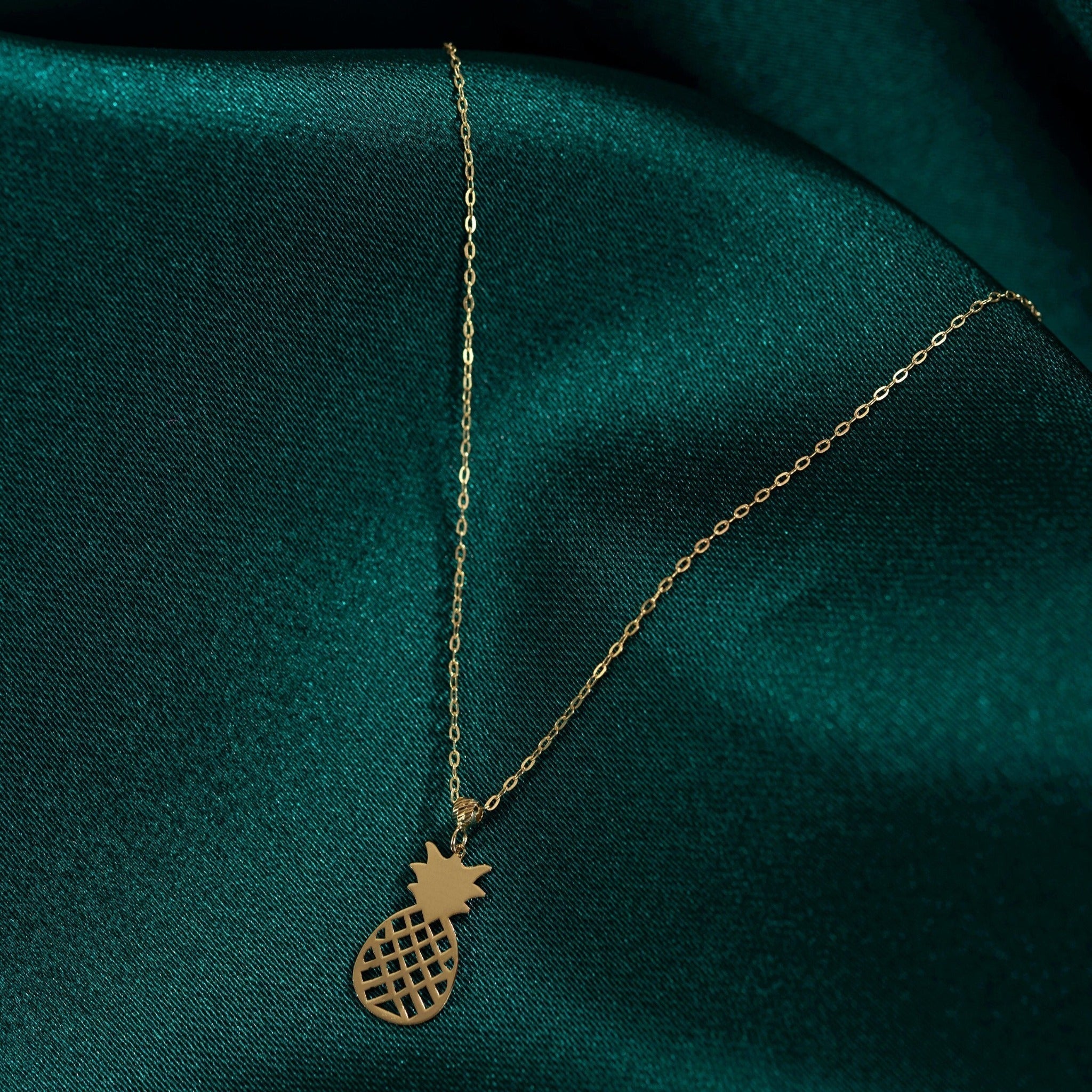 Gold-colored necklace with a fine oval-link chain and open lattice pineapple pendant on dark teal fabric.