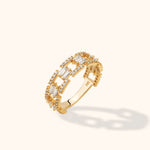 Gold-toned ring with geometric openwork, paired rectangular clear stones and small round stones, angled on a cream background.