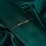 Gold-toned chain bracelet with two infinity links flanking a textured circular ring, displayed on dark teal fabric.
