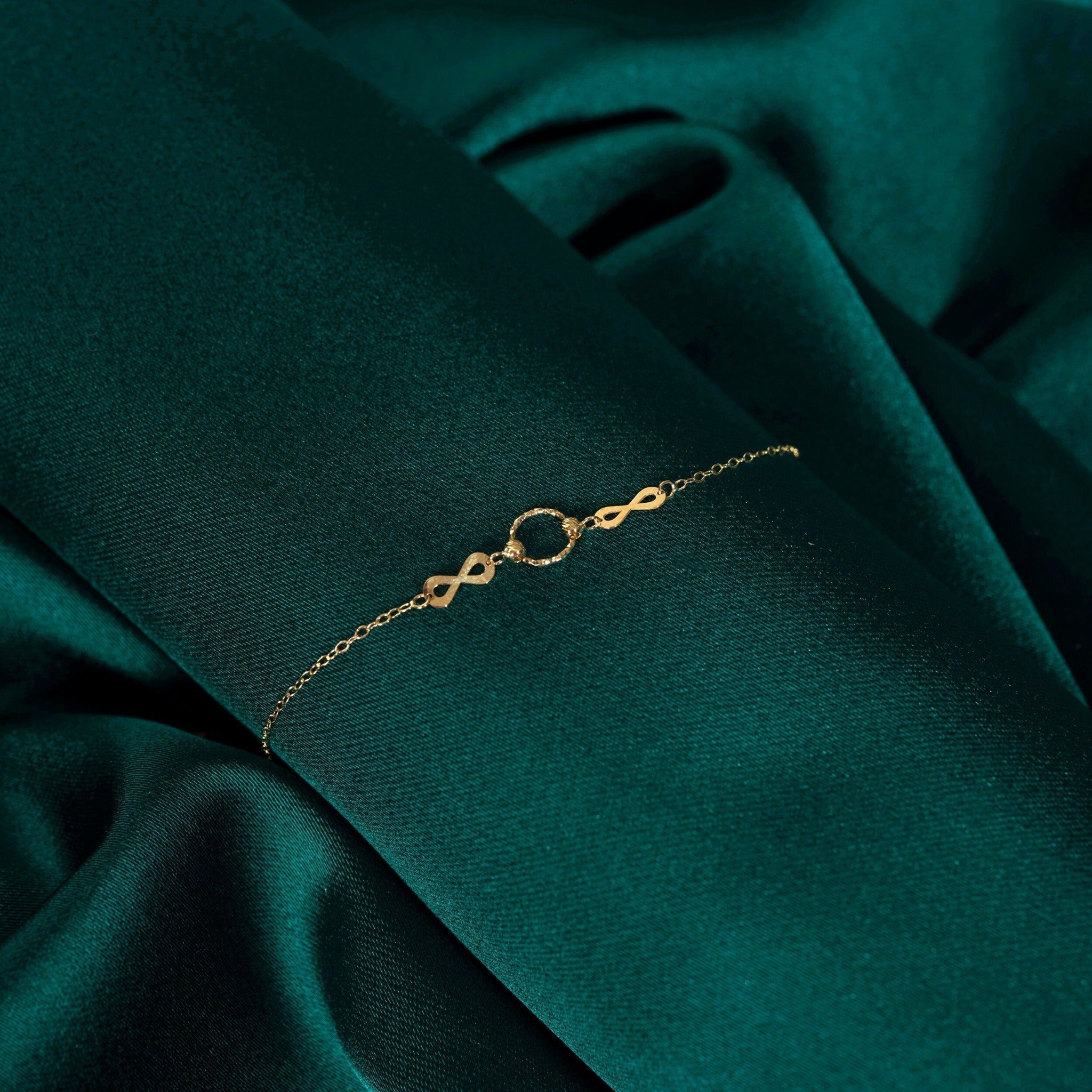 Gold-toned chain bracelet with two infinity links flanking a textured circular ring, displayed on dark teal fabric.