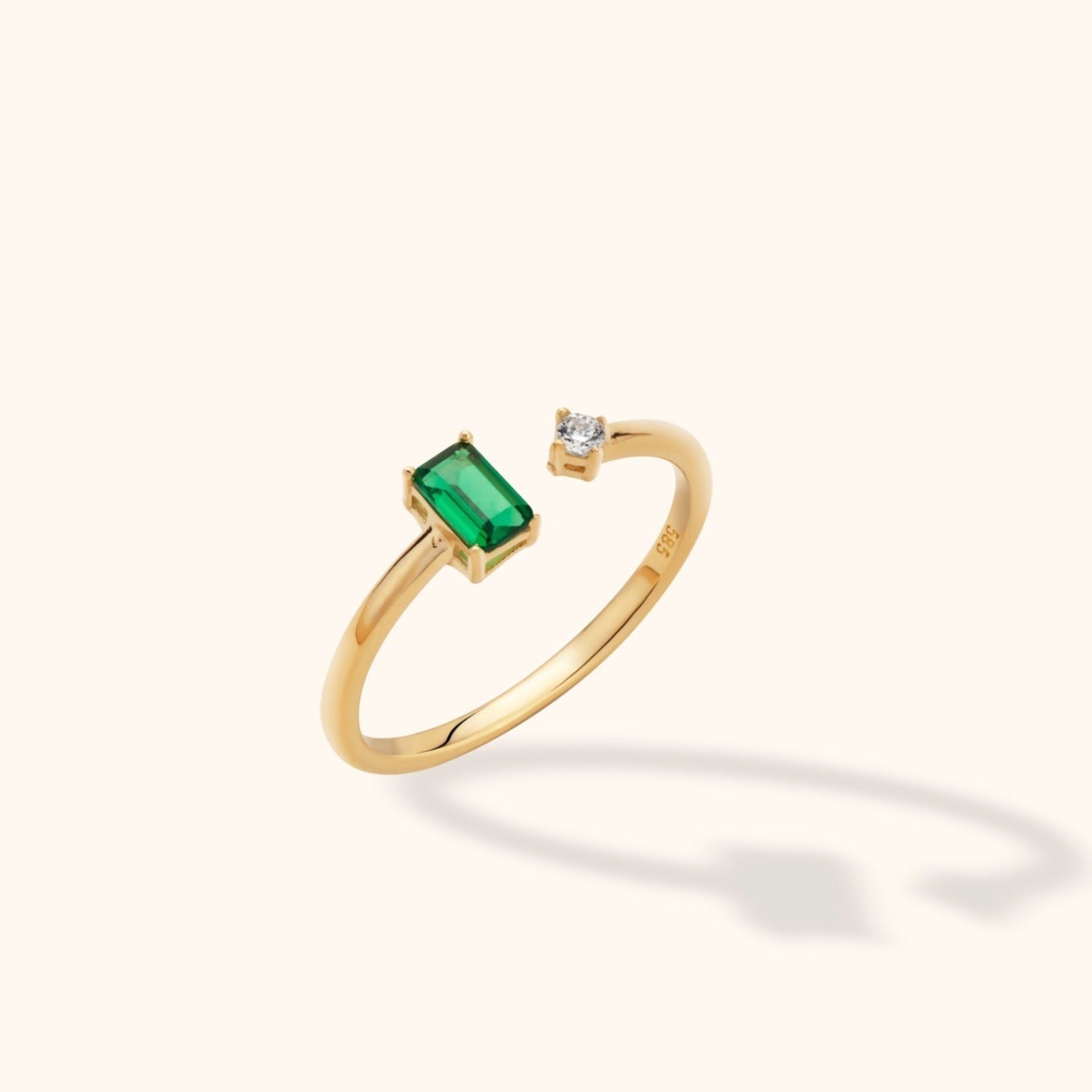 Gold-colored open ring with a rectangular green stone and a small round clear stone at opposite ends of a polished band.