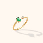 Gold-colored open ring with a rectangular green stone and a small round clear stone at opposite ends of a polished band.