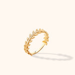 Gold-colored ring with small leaf shapes along both sides of a curved stem, shown tilted on a cream background.