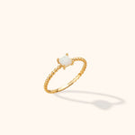 Gold-toned ring with a slim beaded band and a square, milky white center held by four prongs, shown at an angle on a cream background.