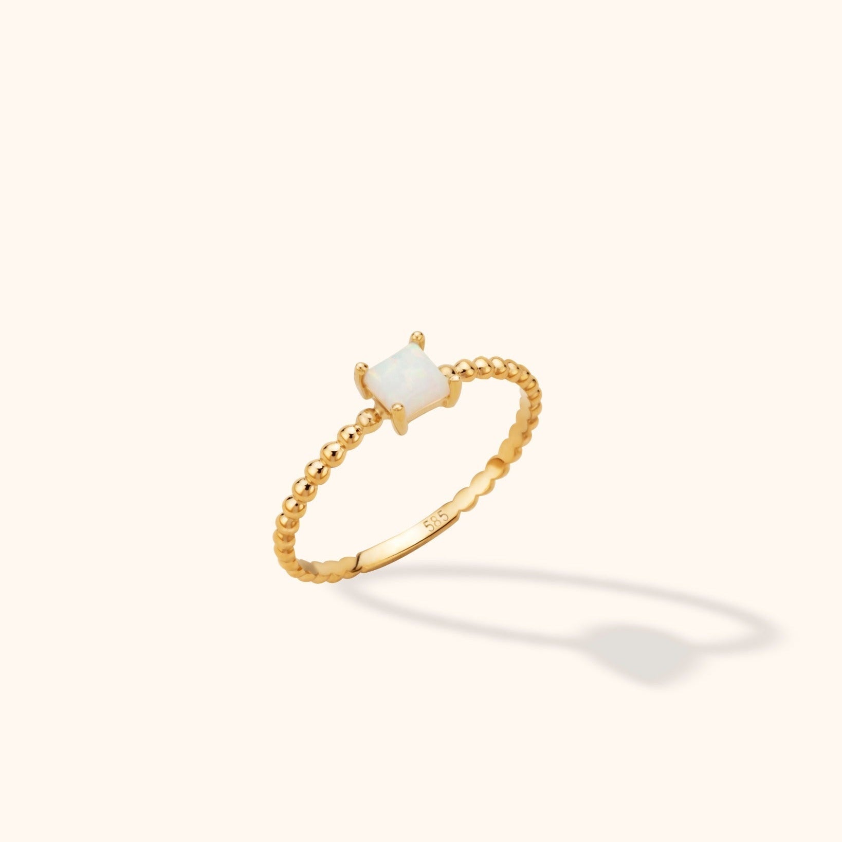 Gold-toned ring with a slim beaded band and a square, milky white center held by four prongs, shown at an angle on a cream background.