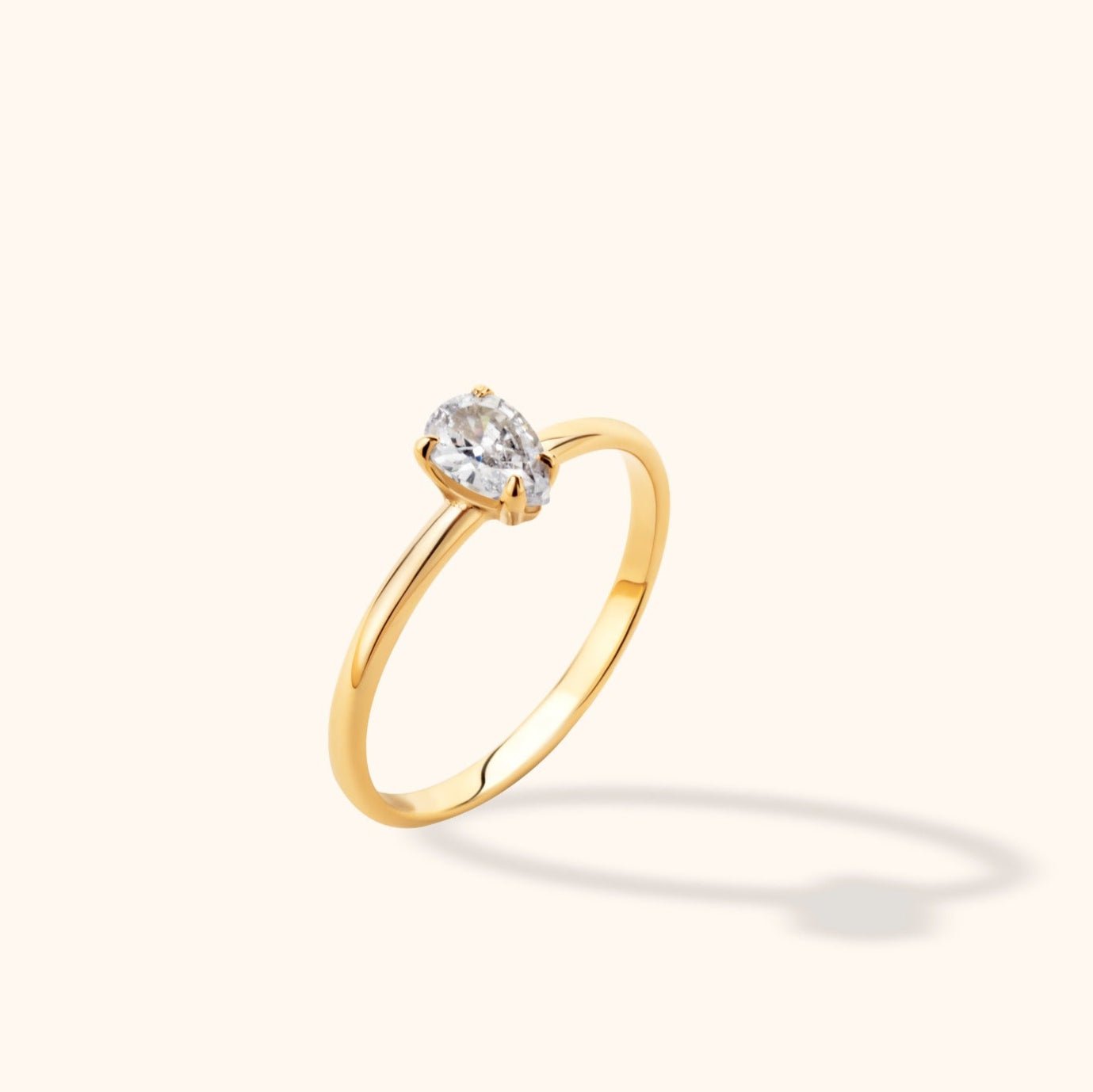 Slender gold-colored ring with a clear faceted stone in a prong setting, shown at an angle against a cream background.